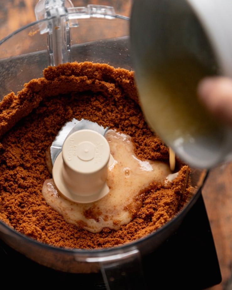 An overhead shot shows melted vegan. butter being poured into a food processor filled with crushed Biscoff cookie crumbs.