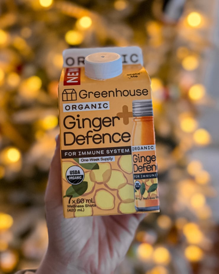 A head-on shot shows a hand holding a carton of Ginger Defense shots against a background of twinkle lights on a Christmas tree.