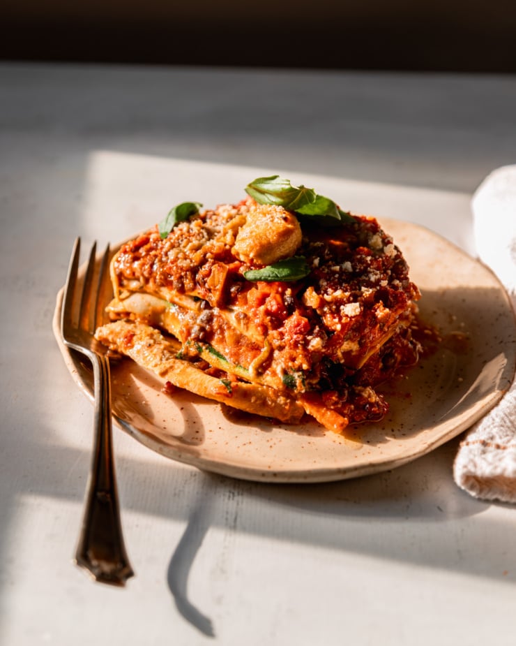 A 3/4 angle shot shows a piece of vegan lasagna on a plate with a fork to the side. The shot is taken in direct sun light.