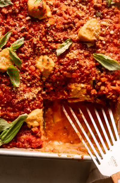 An up close, overhead shot shows a baked vegan lasagna in a baking dish. The top is finished with little dollops of tofu cashew ricotta, fresh basil leaves and vegan "parmesan." One slice has been removed from the dish and a metal spatula rests in the dish.