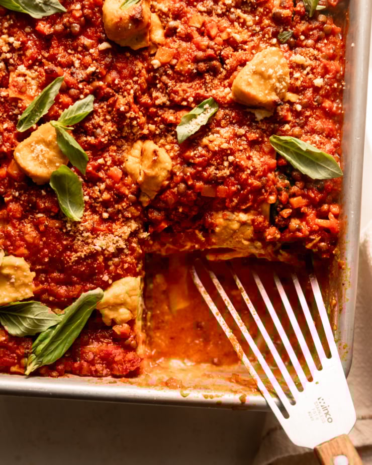 An up close, overhead shot shows a baked vegan lasagna in a baking dish. The top is finished with little dollops of tofu cashew ricotta, fresh basil leaves and vegan "parmesan." One slice has been removed from the dish and a metal spatula rests in the dish.