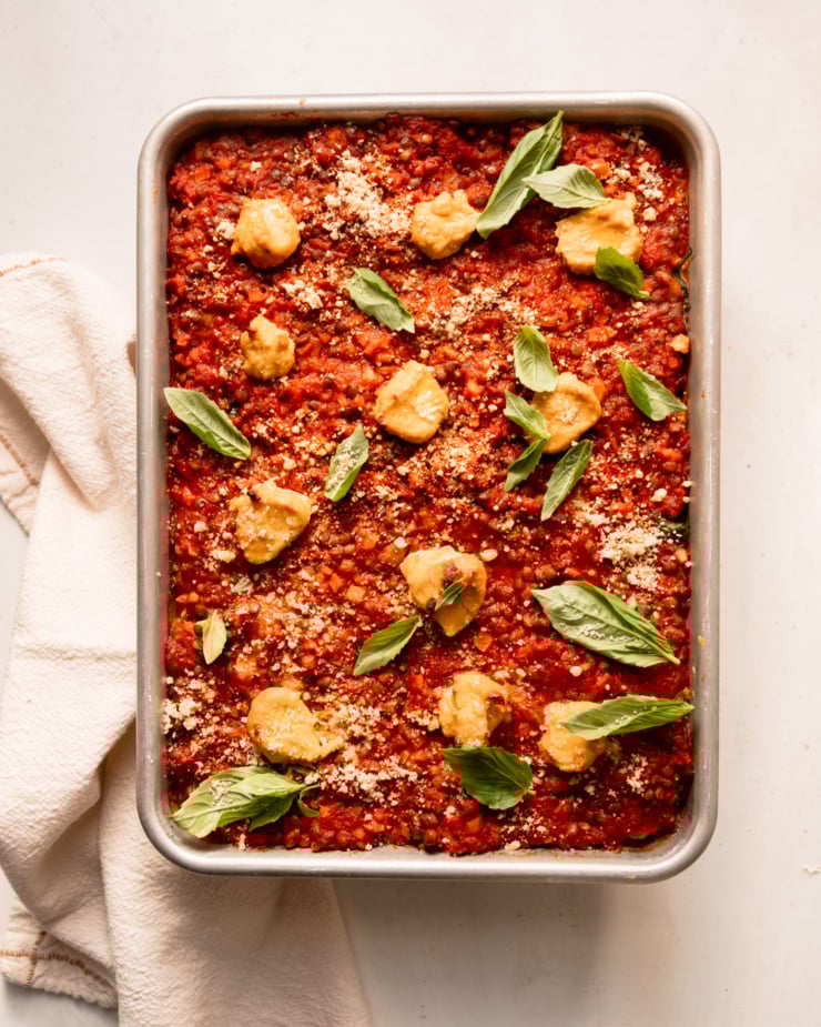 An overhead shot shows a baked vegan lasagna in a baking dish. The top is finished with little dollops of tofu cashew ricotta, fresh basil leaves and vegan "parmesan."