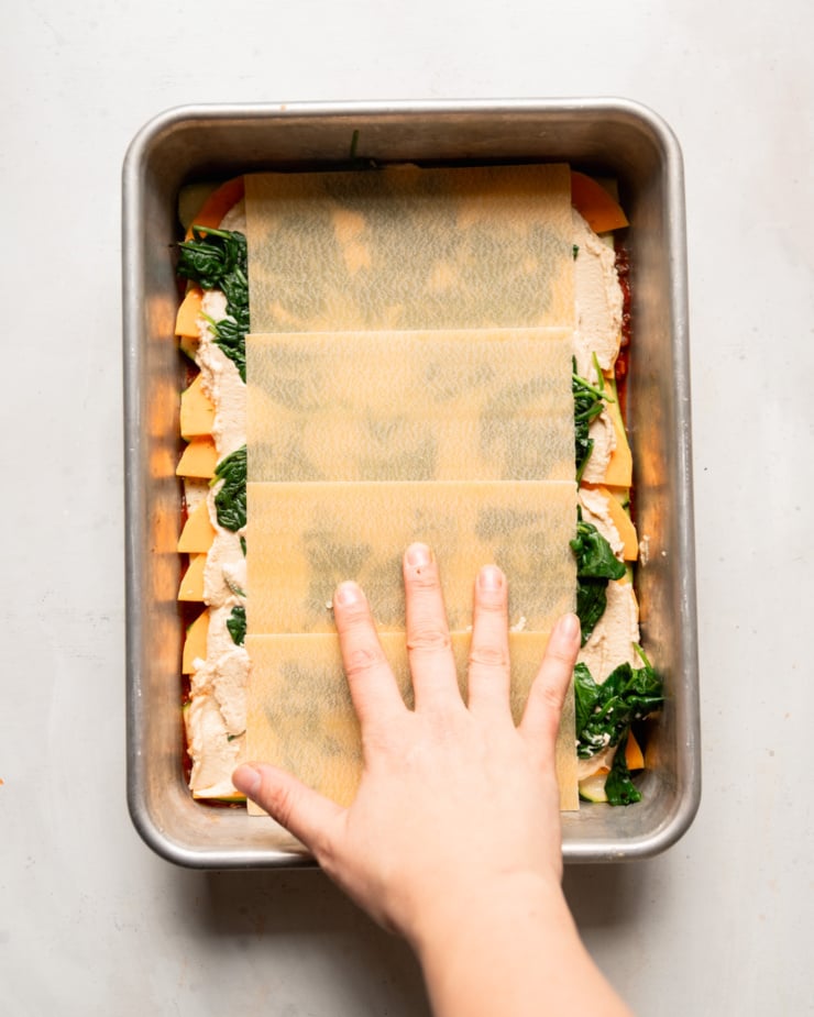 An overhead shot shows a hand pressing lasagna noodles into a baking dish with sliced vegetables and tofu cashew ricotta.