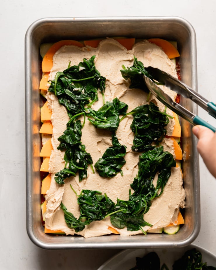 An overhead shot shows a hand using tongs to distribute sautéed baby spinach over a mixture of tofu cashew ricotta and sliced vegetables in a baking dish.