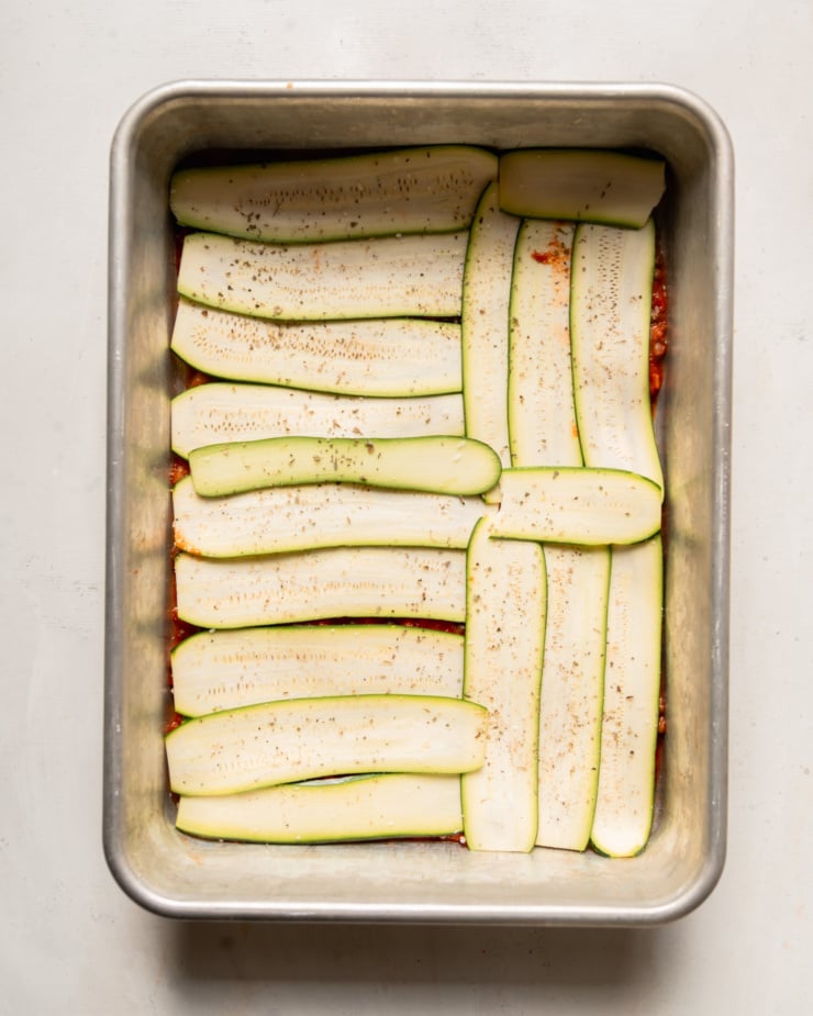 An overhead shot shows thinly sliced zucchini evenly covering a layer of tomato sauce in a baking dish.