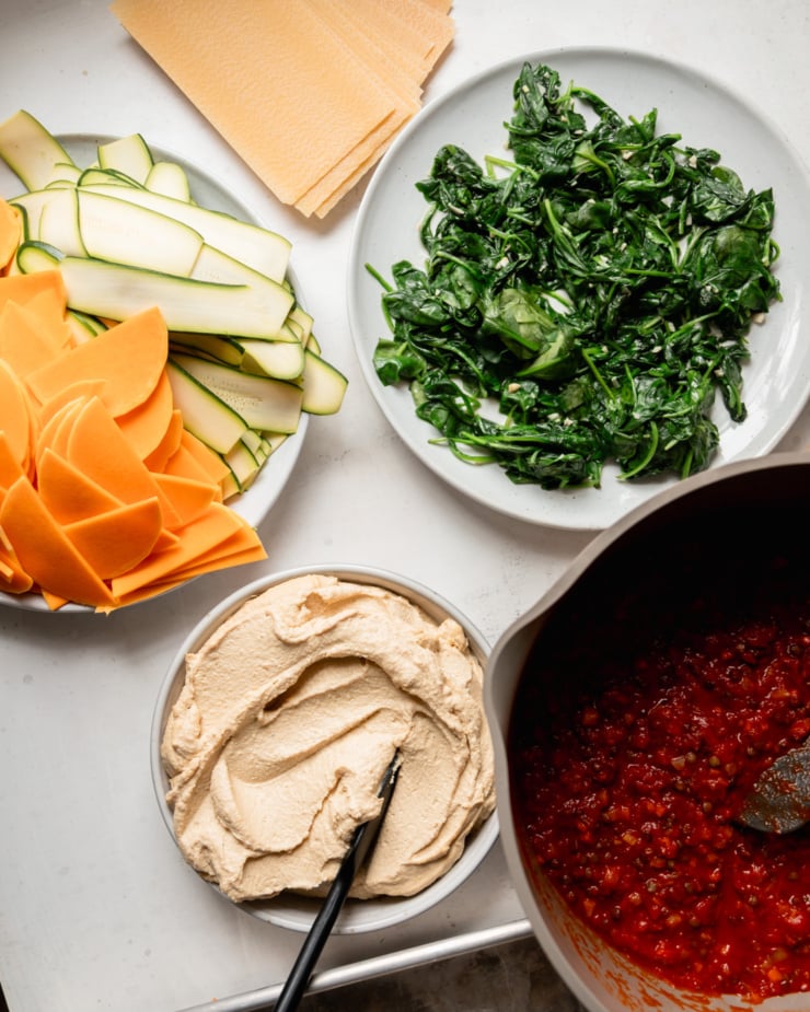 An overhead shot shows components ready to be assembled for a vegan lasagna: oven-ready lasagna noodles, sautéed spinach, lentil and veg-boosted tomato sauce, tofu cashew ricotta, and thinly sliced butternut squash and zucchini.