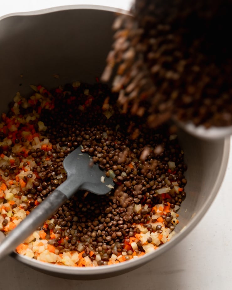 An overhead shot shows a pot filled with sautéed fennel, carrots and shallots. Cooked french lentils are being poured into the pot.