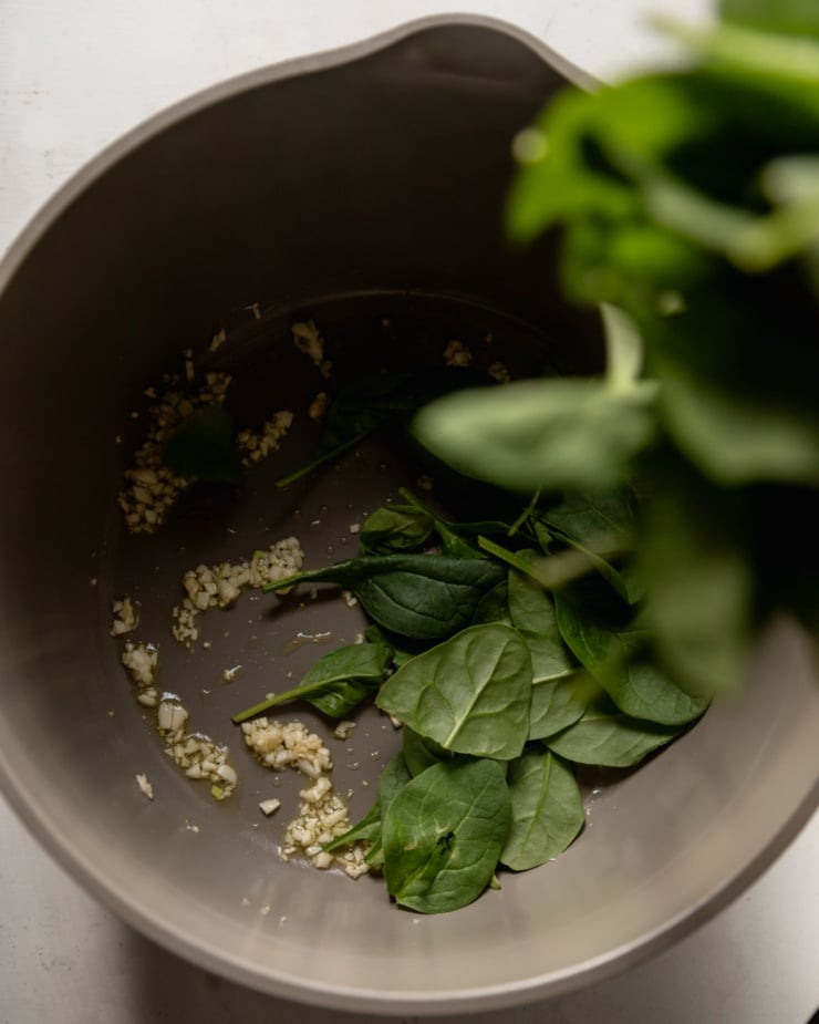 An overhead shot shows a pot with sautéed minced garlic and baby spinach being poured in.
