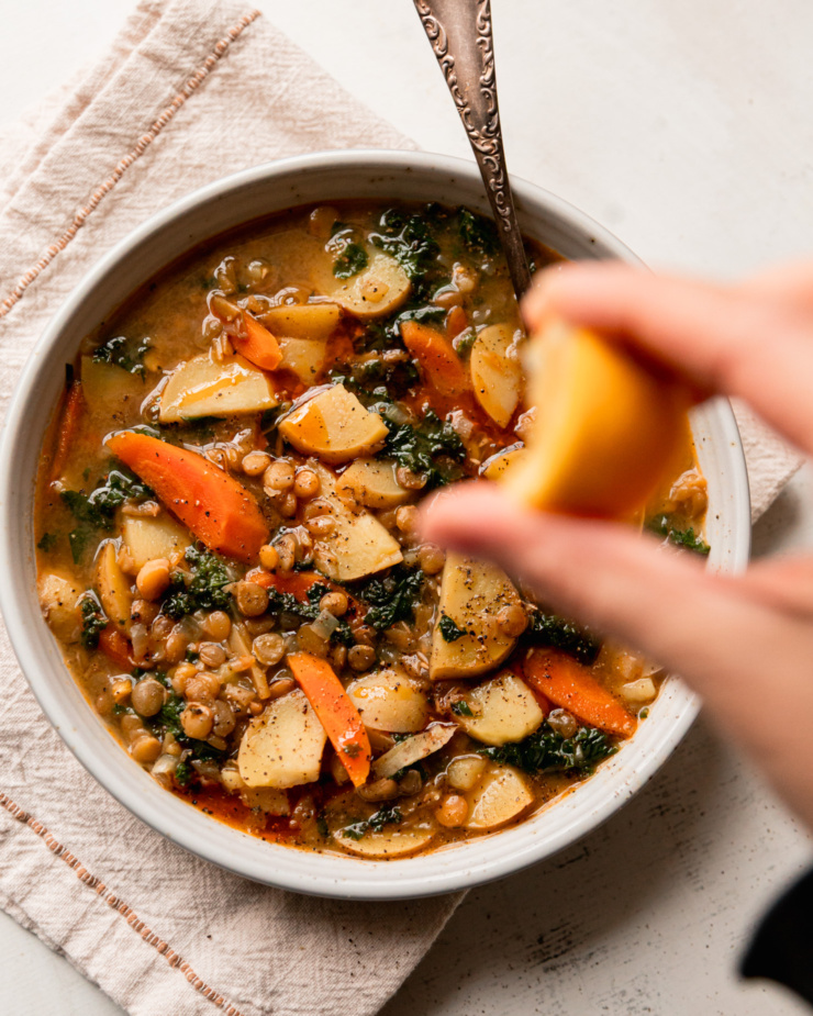 An overhead shot shows a bowl of lentils and potatoes soup with a hand squeezing a lemon wedge over the top.