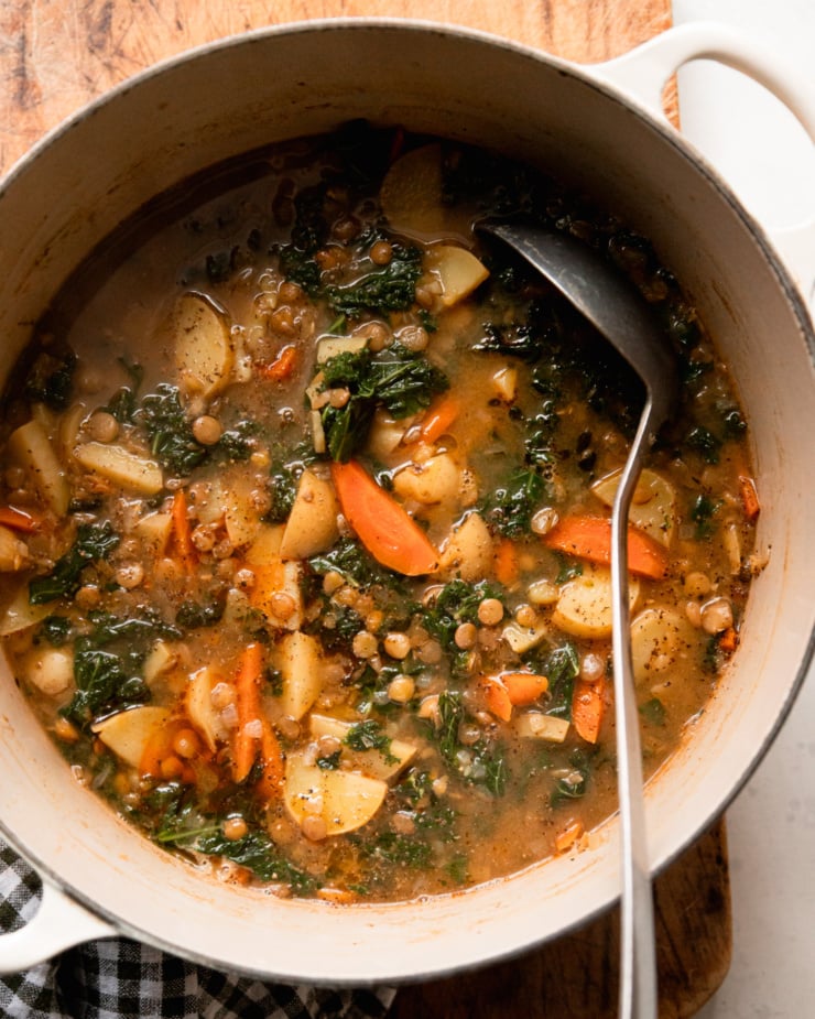 An overhead shot shows a Dutch oven filled with lentils and potatoes soup with greens and carrots. A ladle is in the pot.