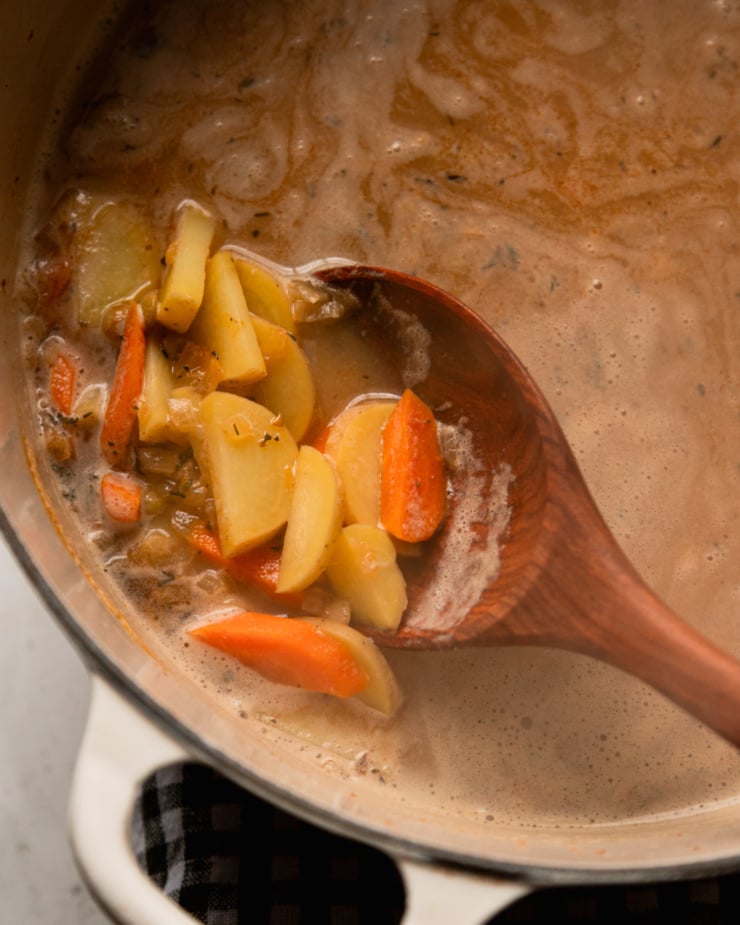 An overhead shot shows a wooden spoon pulling up some cooked carrots and potatoes from a pot of brothy soup.