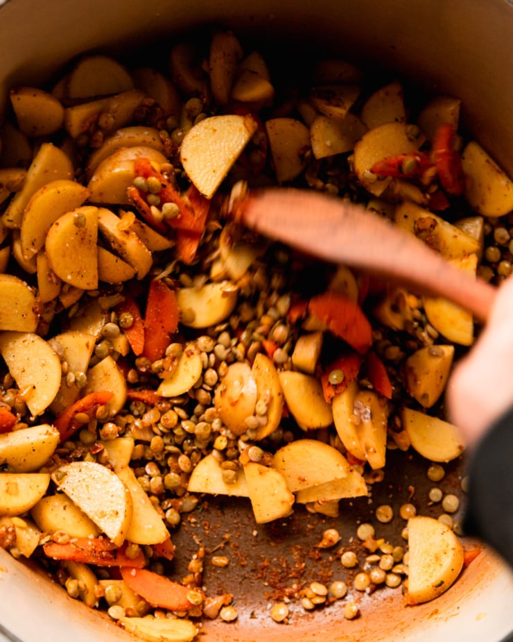 An overhead shot shows a hand using a wooden spoon to stir up carrots, potatoes, green lentils, and spices.