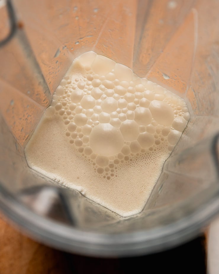 An overhead shot shows a blended mixture of vegetable stock, roasted garlic cloves, and miso in a blender pitcher. It is bubbly and foamy at the top.