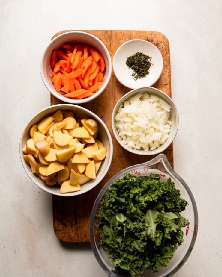 An overhead shot shows bowls containing: sliced carrots, minced thyme, diced onion, chopped kale, and sliced potatoes.