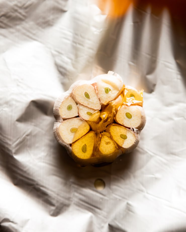An overhead shot shows a head of garlic with the top third cut off. It is sitting on top of a sheet of foil and olive oil has been drizzled on top.