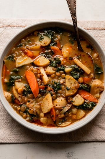 An overhead shot shows a bowl of lentils and potatoes soup with carrots, kale, and roasted garlic broth. A spoon is sticking out of the bowl and black pepper has been ground on top. Little dabs of chili oil can also be seen on the surface of the soup.