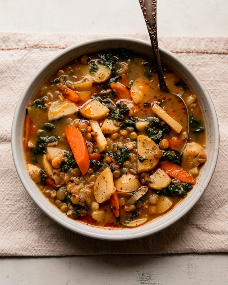 An overhead shot shows a bowl of lentils and potatoes soup with carrots, kale, and roasted garlic broth. A spoon is sticking out of the bowl and black pepper has been ground on top. Little dabs of chili oil can also be seen on the surface of the soup.