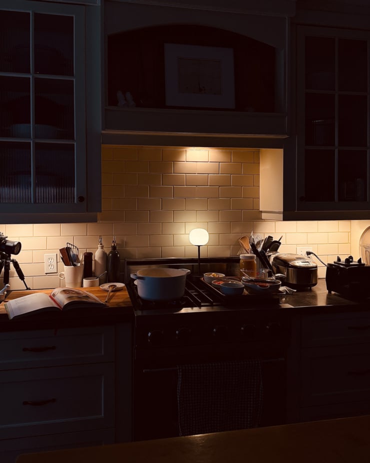A head on shot shows a kitchen stove and counter area in low lighting, all provided from under the cabinets. A pot, cookbook, and a tray with prepped ingredients are all in the scene.