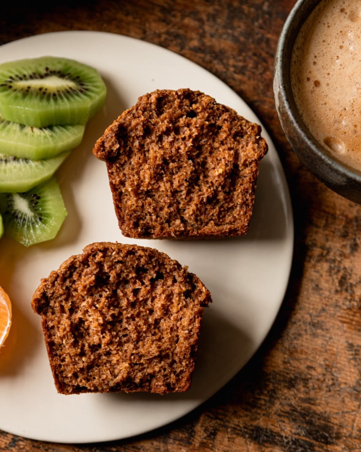 An up close, overhead shot shows a split vegan bran muffin on a plate alongside some sliced kiwi.