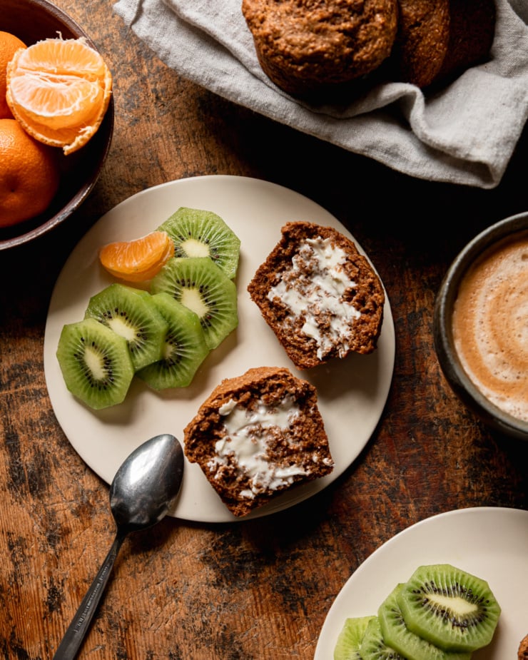 An overhead shot shows a split vegan bran muffin on a plate next to some sliced kiwi and peeled clementine. The muffin has vegan butter spread on both sides. A coffee and more fruit is nearby.
