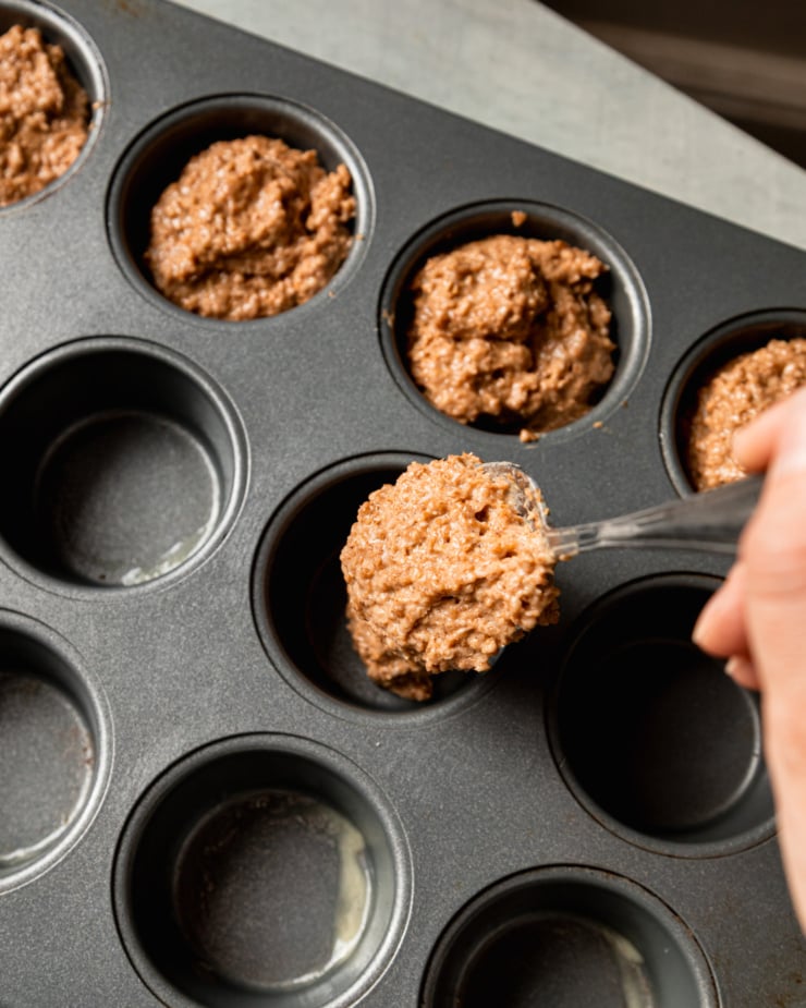 An overhead shot shows a hand using a spoon to portion batter into a muffin tin.