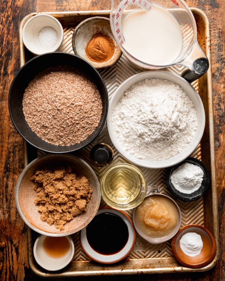 An overhead shot shows measured ingredients in prep bowls and measuring cups for a batch of vegan bran muffins. All bowls are on a baking sheet.