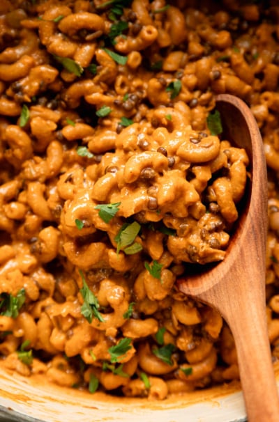 An up close, overhead shot shows a vegan hamburger helper-style mixture with lentils, elbow macaroni, and chopped parsley. A wooden spoon is sticking out of the mixture.