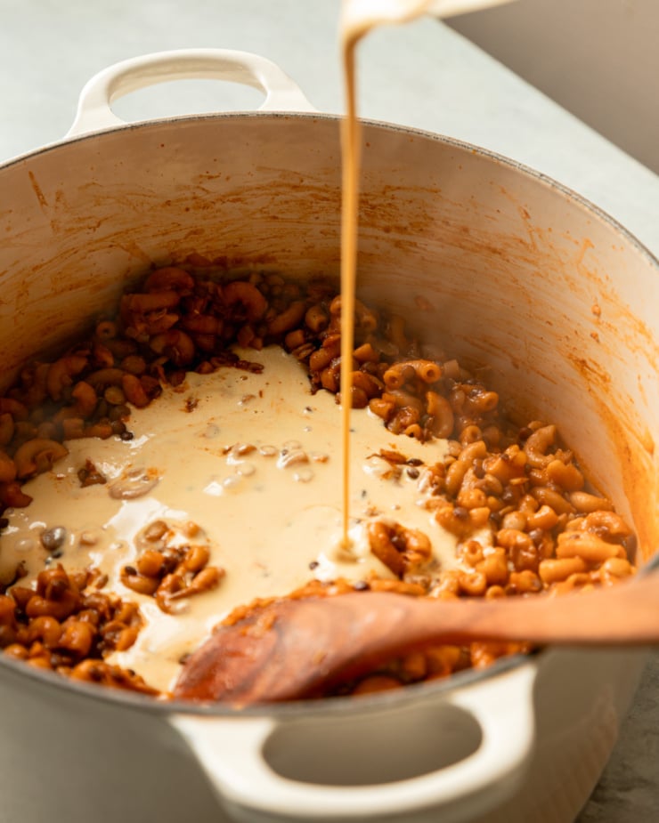A 3/4 angle shot shows a savory vegan "cream" being poured into a pot with pasta and lentils.