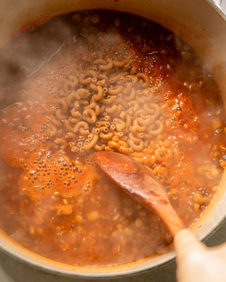 An overhead shot shows a tomato-y brothy mixture in a pot with whole wheat elbow macaroni poured on top. There is lots of steam rising from the pot.