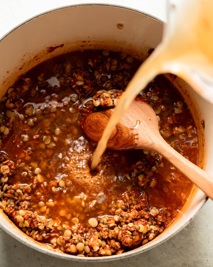 An overhead shot shows vegetable stock being poured into a pot with a spiced, tomato-y lentil mixture. A wooden spoon is also sticking out of the pot.