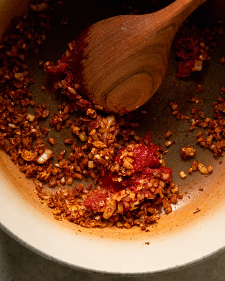 An overhead shot shows diced onions and spices that have been sautéed along with tomato paste in a pot. A wooden spoon is stirring everything together.