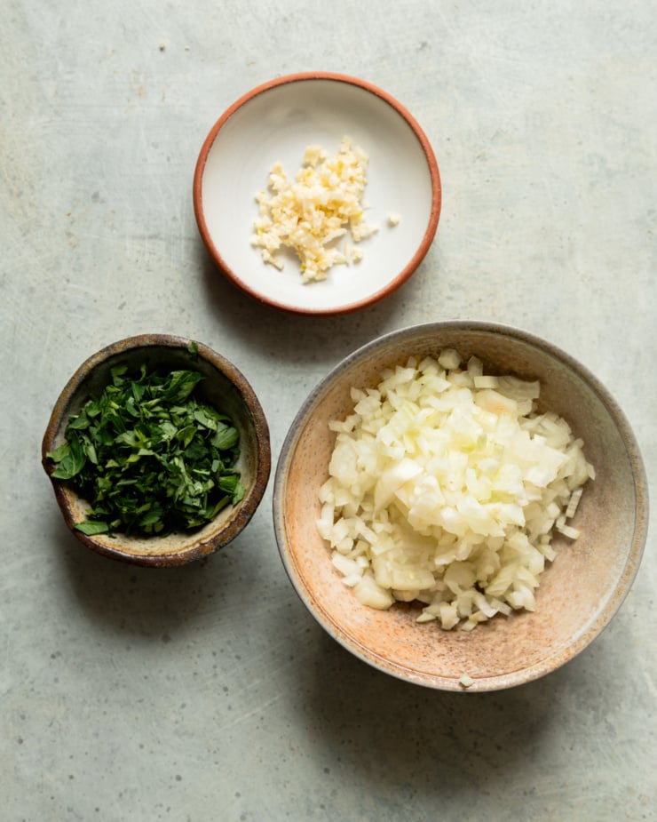 An overhead shot shows 3 bowls filled with small diced onion, minced garlic, and chopped parsley.