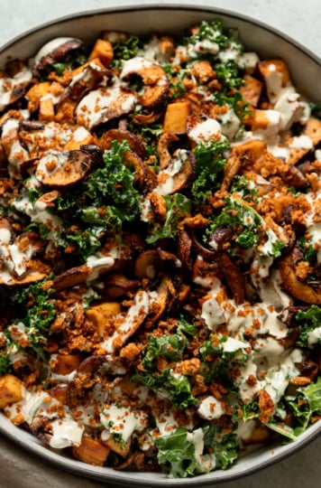 An up close, overhead shot shows a wide dish filled with roasted mushrooms and kale salad. The salad is topped with creamy garlic cashew dressing and seasoned bread crumbs