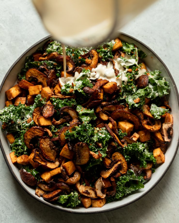 An overhead shot shows creamy garlic cashew dressing being poured over dressed chopped kale, roasted mushrooms, and smoked tofu.