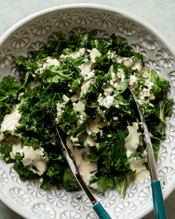 An overhead shot shows chopped kale in a bowl that has been drizzled with creamy garlic cashew dressing. A pair of tongs sticks out of the bowl.