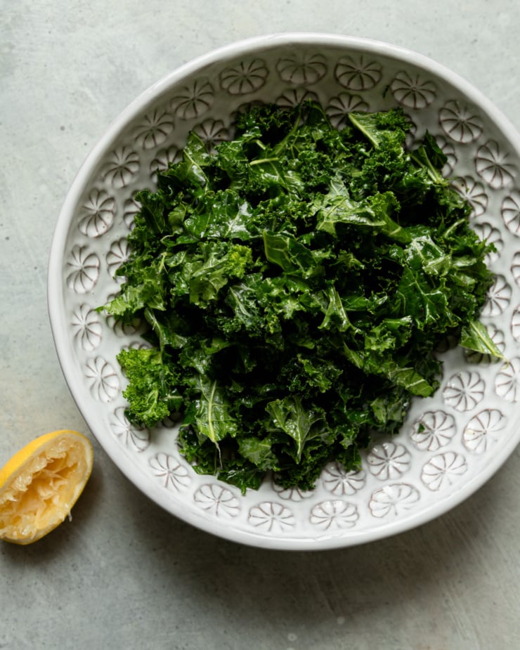 An overhead shot shows a bowl filled with chopped kale that has been massaged with lemon juice and olive oil. A squeezed lemon half is seen to the side.