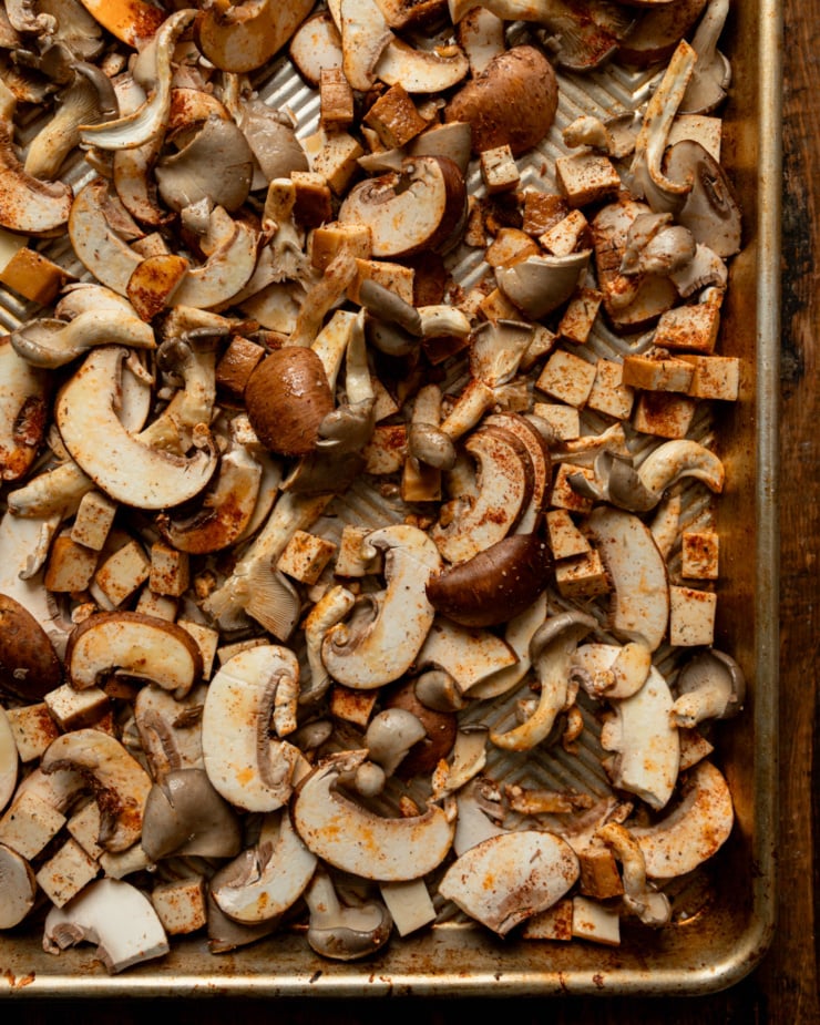 An overhead, up close shot shows sliced mushrooms and cubes of smoked tofu dusted with spices on a baking sheet.