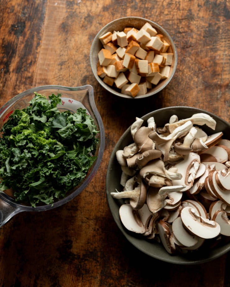 An overhead shot shows prepped ingredients in 3 containers: diced smoked tofu, sliced mushrooms, and chopped kale.