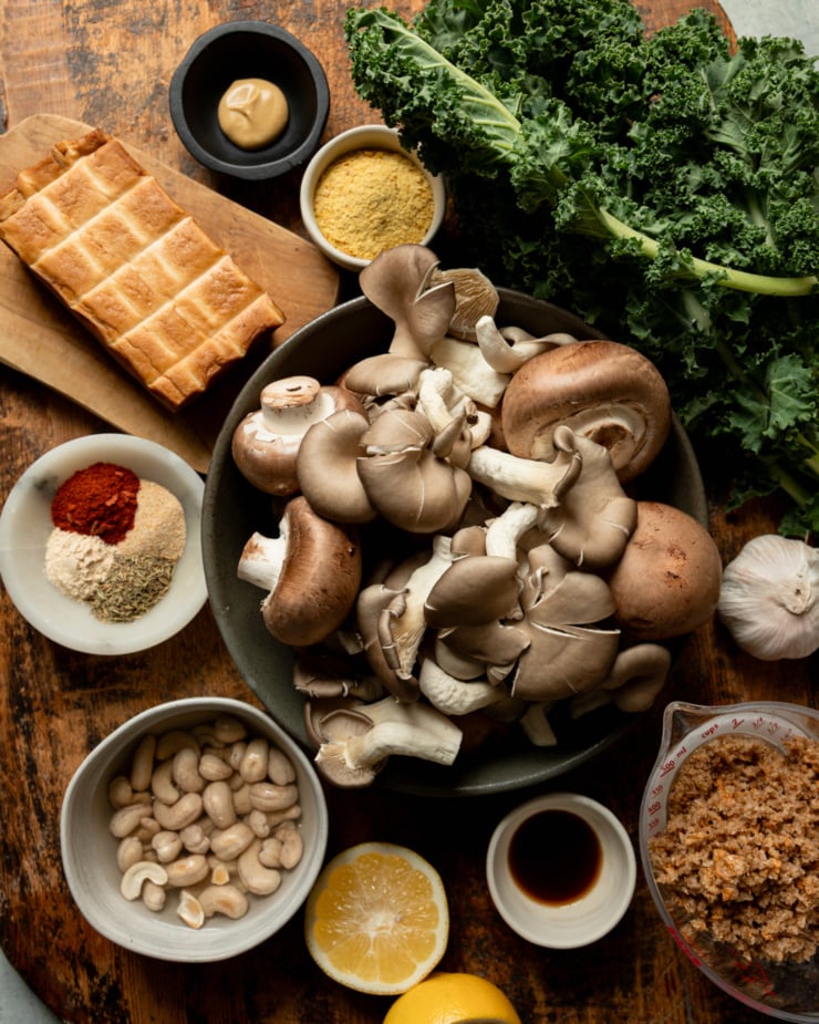 An overhead shot shows ingredients needed for a roasted mushroom and tofu kale salad. All ingredients are on a rough wooden board in natural lighting.