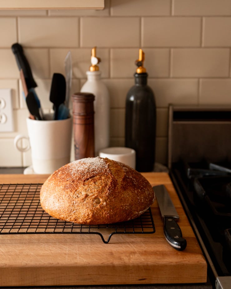 A head-on shot shows a loaf of homemade whole wheat bread on a cooling rack next to a stove. Bottles of oil, salt, pepper mill, and a carafe of utensils is seen in the background. A serrated knife is next to the bread on a cooling rack.