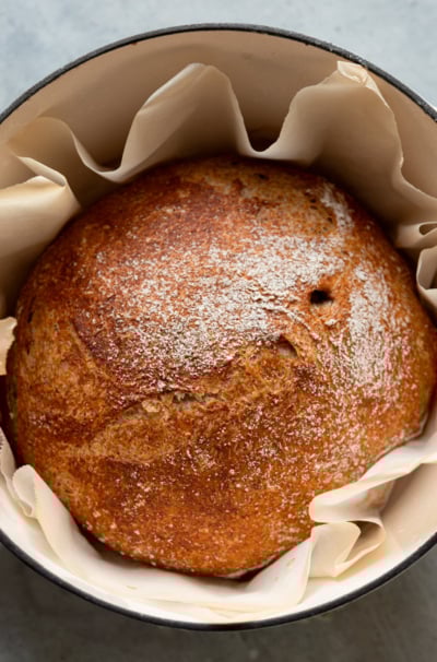 An overhead shot shows a baked round loaf of no-knead whole wheat bread in a Dutch oven, surrounded by parchment paper.
