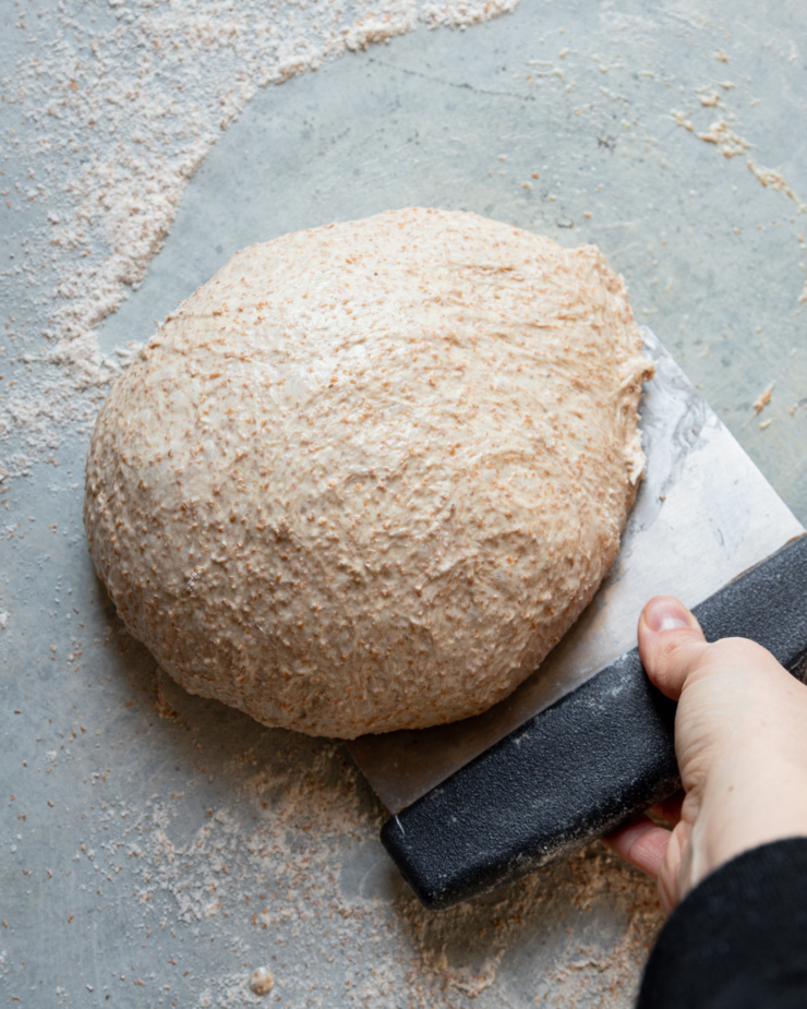 An overhead shot shows a hand using a bench scraper to shape bread dough into a round loaf.
