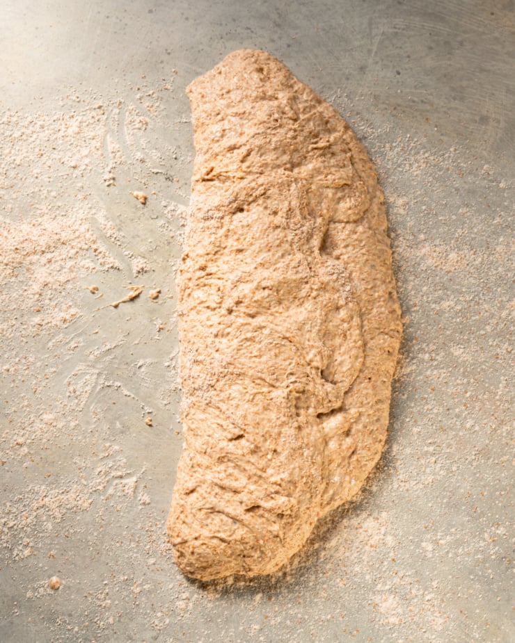 An overhead shot shows a long piece of bread dough that has been folded in half on a floured surface.